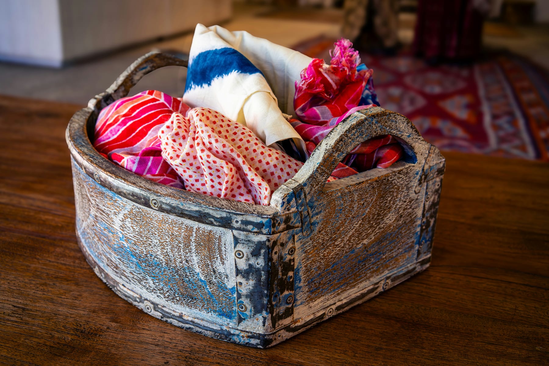A worn wooden basket on a table, filled with brightly patterned folded cloth