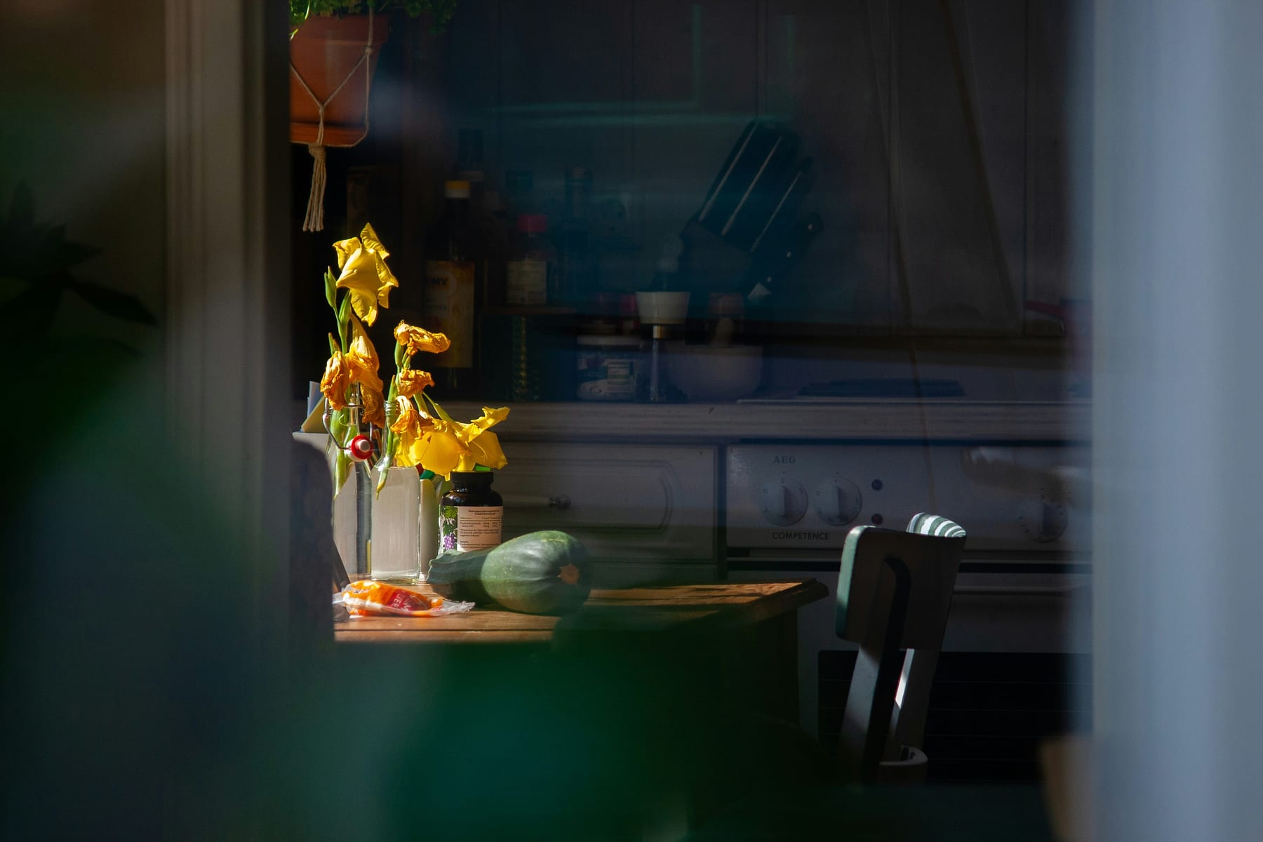 A kitchen table seen through a window, daffodils in a jar and vegetables on a board
