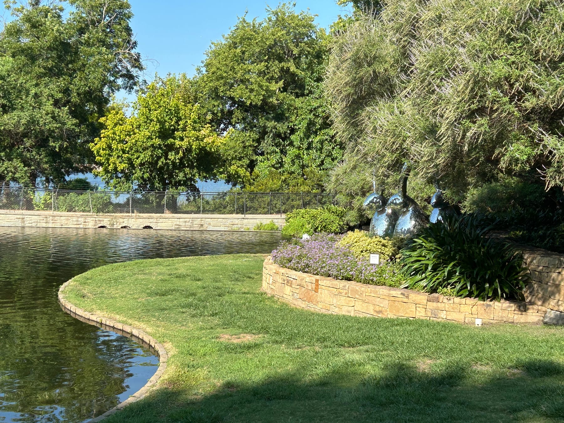 A lawn curving around the pond at the Arboretum, with a stone planter bed and chrome bird sculptures under the trees