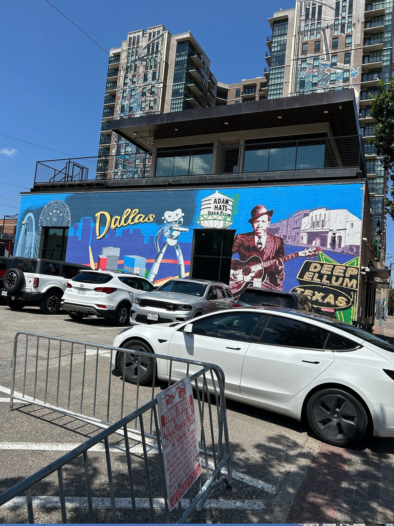 The Deep Ellum mural on a brick wall - a guitarist in a hat, the Dallas skyline, and the Adam Hats sign above the parked cars