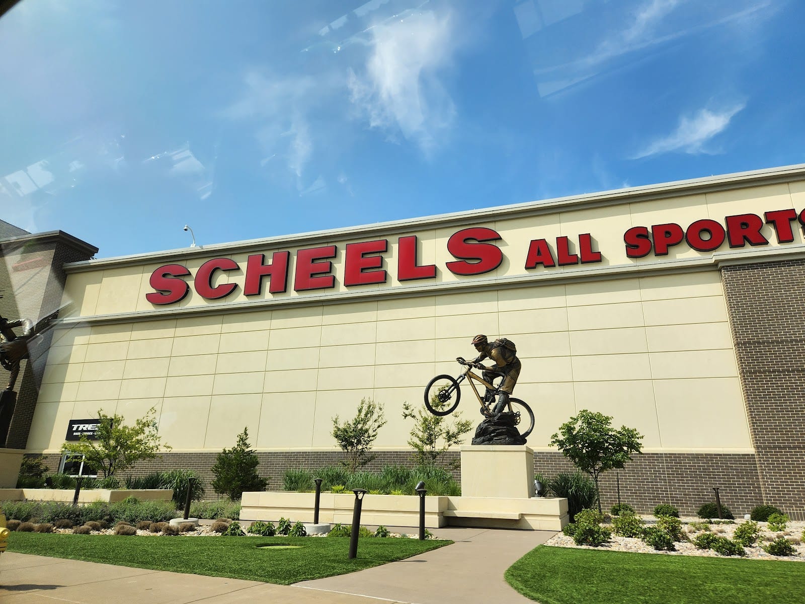 The Scheels All Sports sign across the front of the building against a blue sky, with a bronze statue of a mountain biker on a plinth below it.